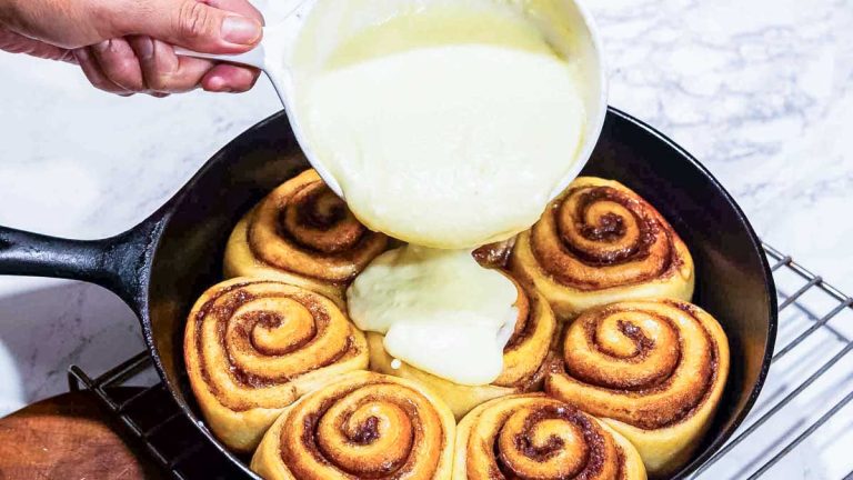 one hand pouring frosting over sourdough brioche cinnamon rolls in a cast iron pan