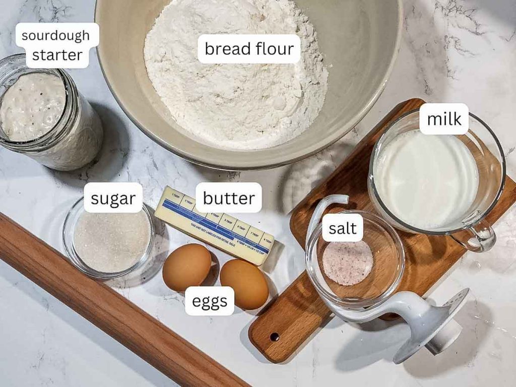 sourdough brioche cinnamon rolls ingredients; flour, sourdough starter inside jar, milk in a cup, butter, eggs, salt, sugar with rolling pin and chopping board on surface