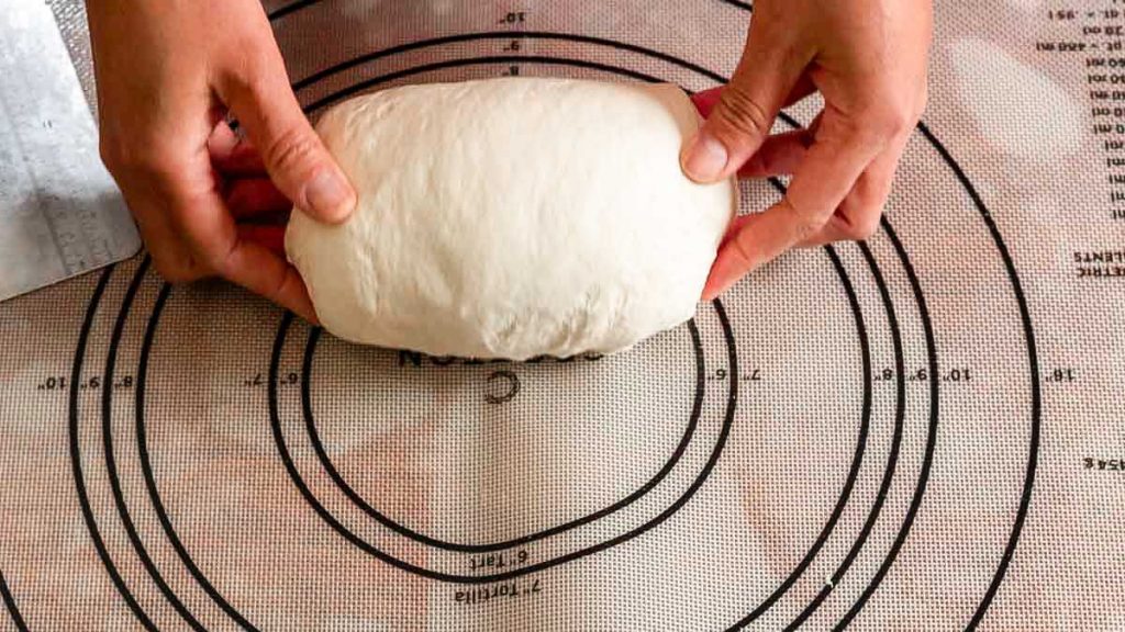 two hands rolling sourdough sandwich wet dough into a loaf on a mat step 3